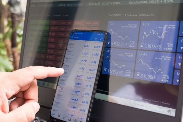 En smartphone med en Forex trading app öppnad på skärmen, som lutar mot en dator skärm som också visar valuta kurser och trading.